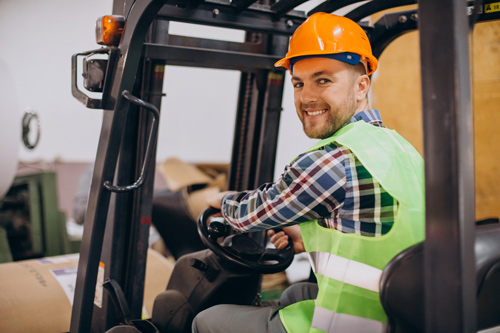 A smiling man in a hardhat and flanel shirt operates a forklift.